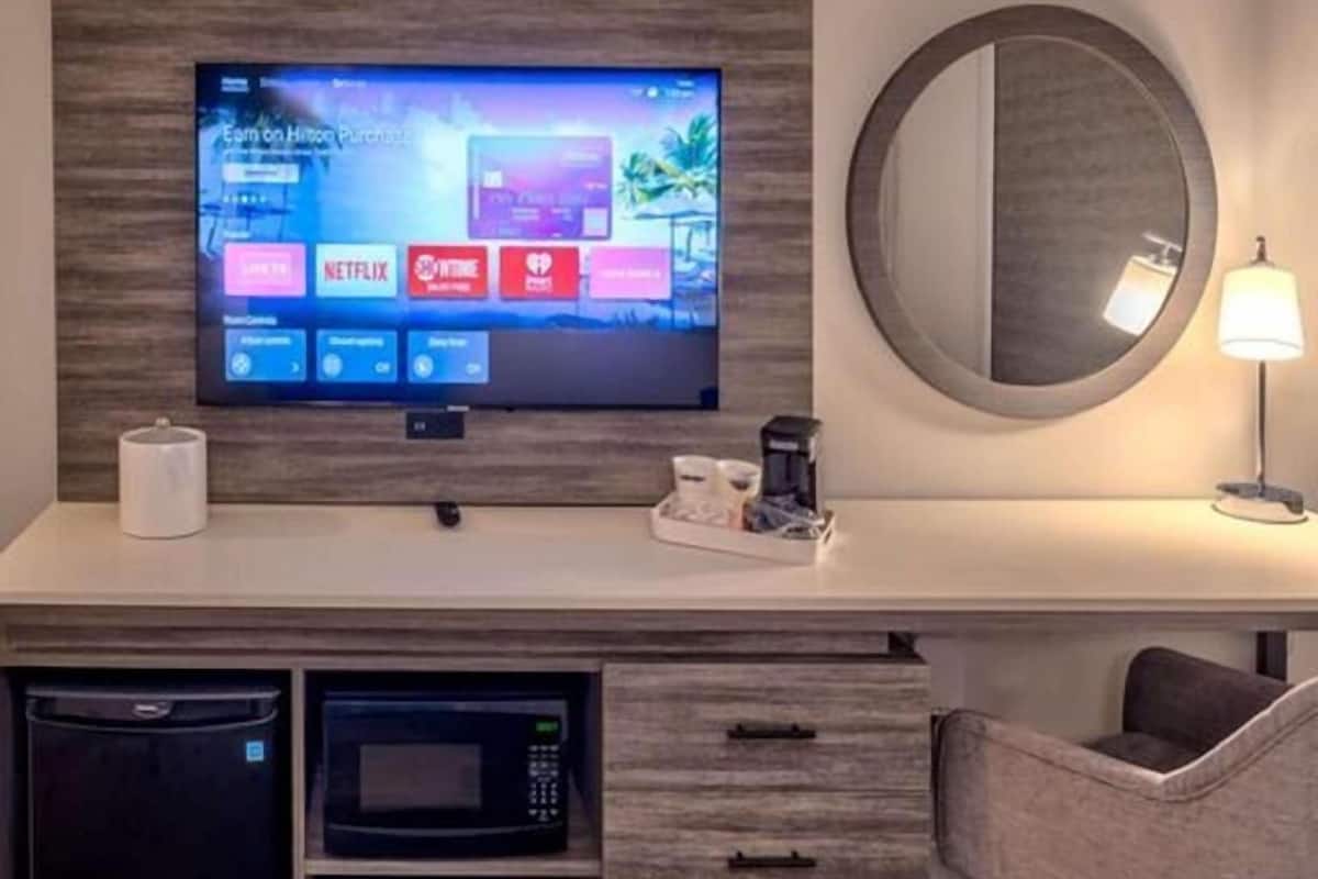Une chambre d’hôtel équipée d’un téléviseur à écran plat permettant d’accéder aux services de diffusion en continu, d’un four à micro-ondes, d’une machine à café, d’un miroir rond et d’un lit recouvert d’une couette blanche.