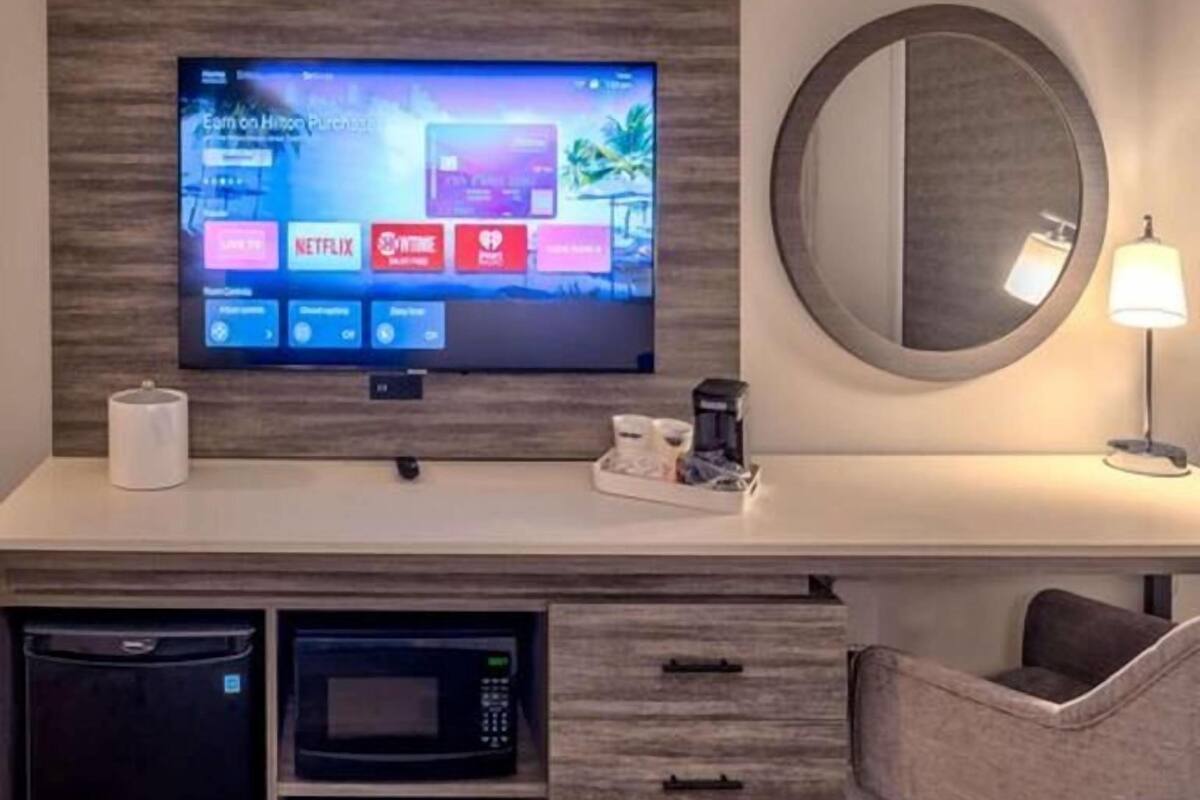 Une chambre d’hôtel équipée d’un téléviseur à écran plat permettant d’accéder aux services de diffusion en continu, d’un four à micro-ondes, d’une machine à café, d’un miroir rond et d’un lit recouvert d’une couette blanche.