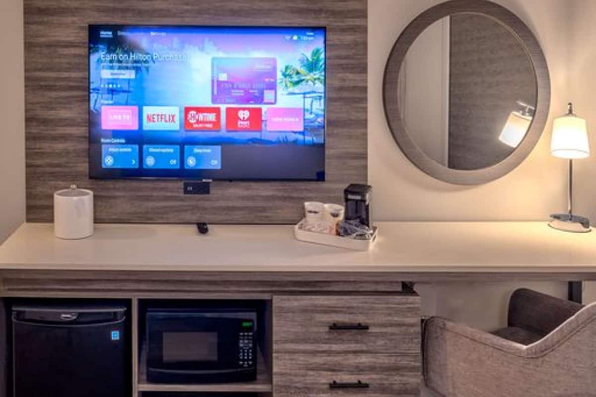 Una habitación de hotel con televisión de pantalla plana montada en una pared de madera, un espejo redondo, una mesita de noche con lámpara, un microondas y una cafetera.