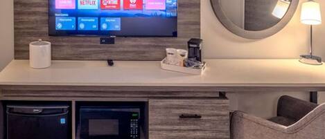 Une chambre d’hôtel dotée d’un téléviseur à écran plat fixé à un mur en bois, d’un miroir rond, d’une table de chevet avec une lampe, d’un four à micro-ondes et d’une machine à café.