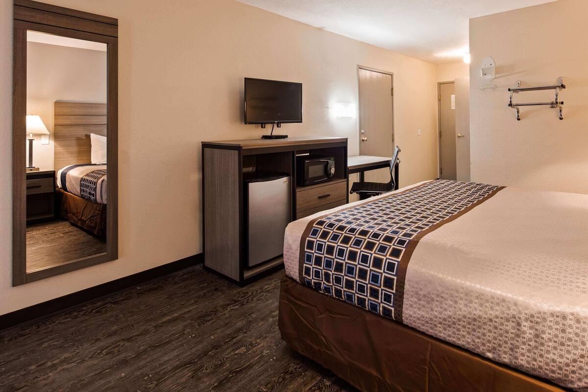 A hotel room with a bed, a desk with a TV, a mirror, and a door.