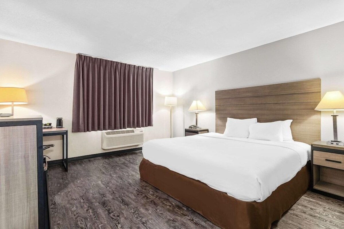 A hotel room with a large bed, two bedside tables with lamps, a wooden headboard, a window with curtains, and a built-in air conditioning unit.