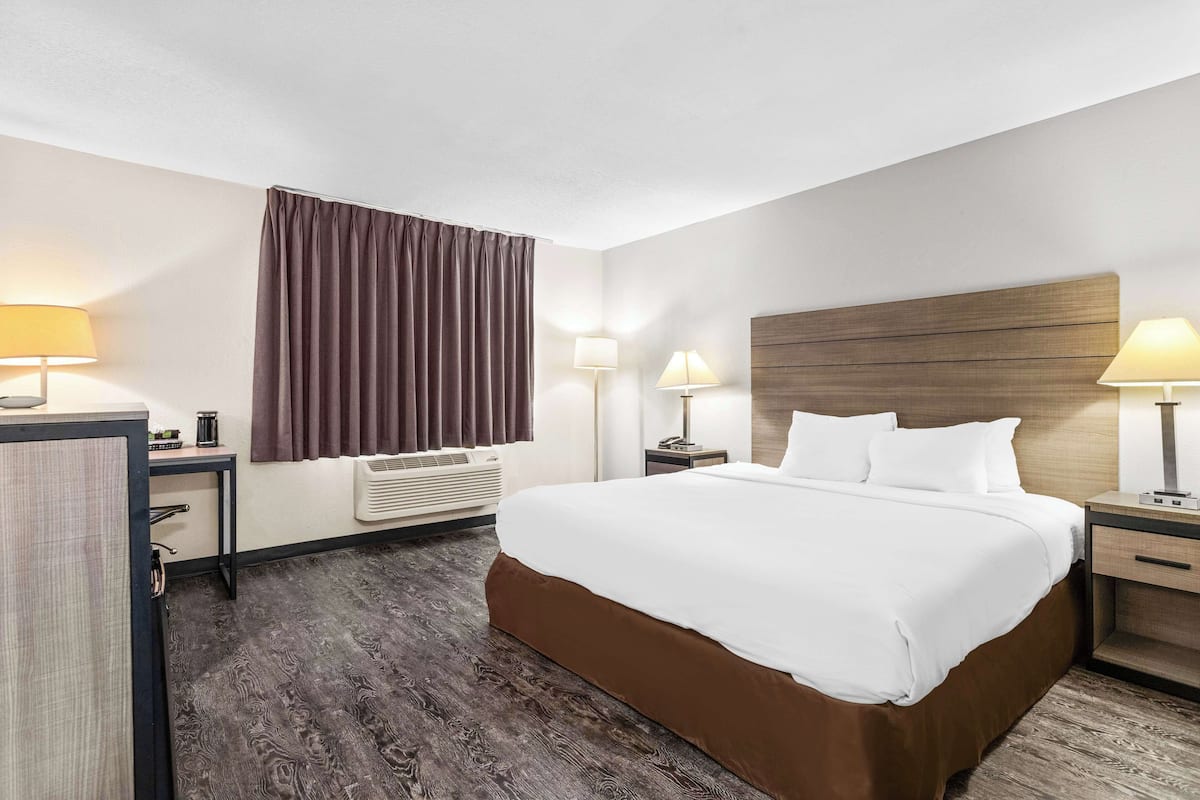 A hotel room with a large bed, two bedside tables with lamps, a window with curtains, and a wooden headboard.