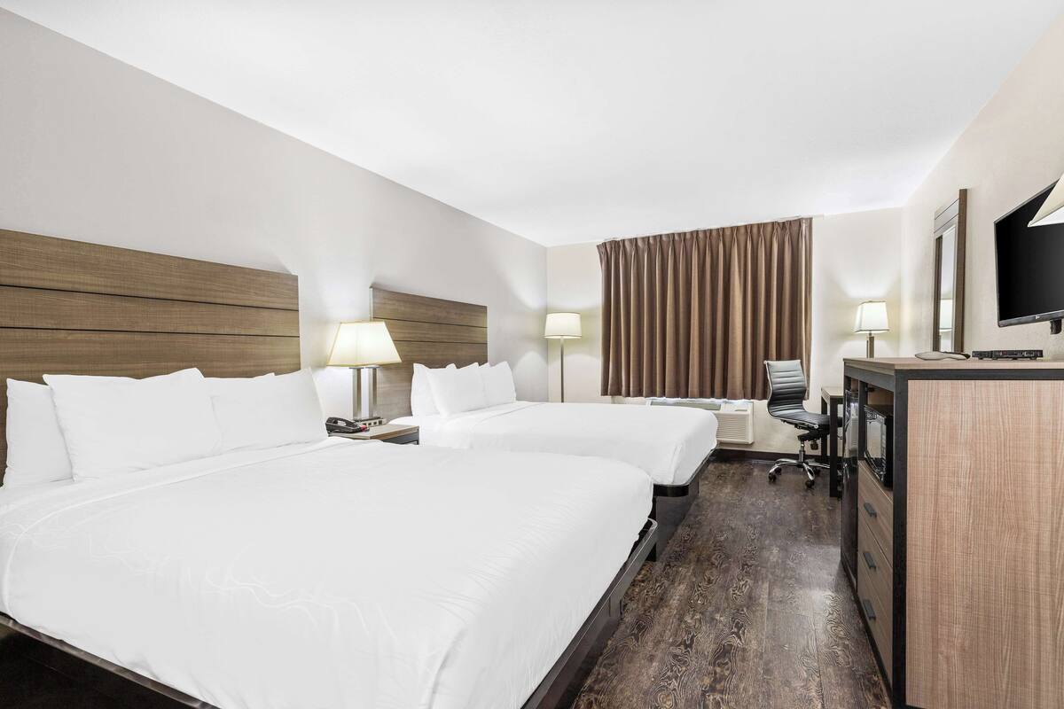 A hotel room with two beds, a desk with a chair, a TV, and a large window with curtains.