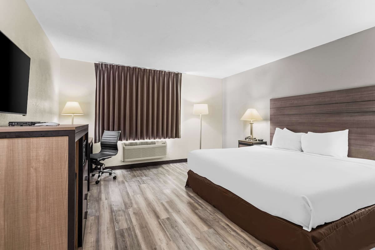 A hotel room with a large bed, a desk with a chair, a flat-screen TV, two bedside lamps, and a window with curtains.