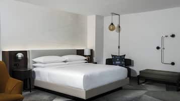 A modern hotel room with a large bed, a bedside table with a lamp, a bench, and a wall-mounted light fixture.