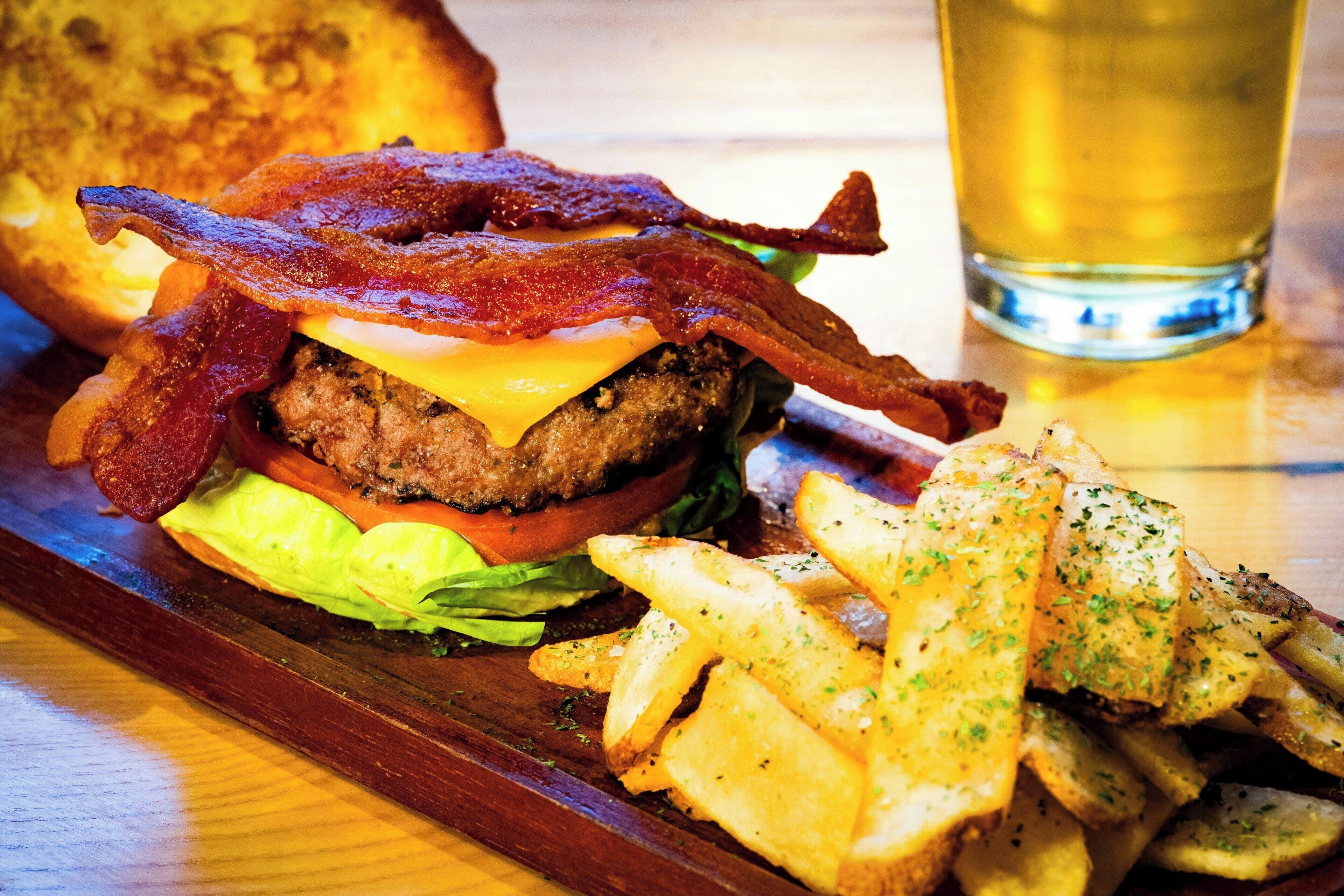 A burger with bacon, cheese, and lettuce, served with fries and a glass of beer on a wooden table.