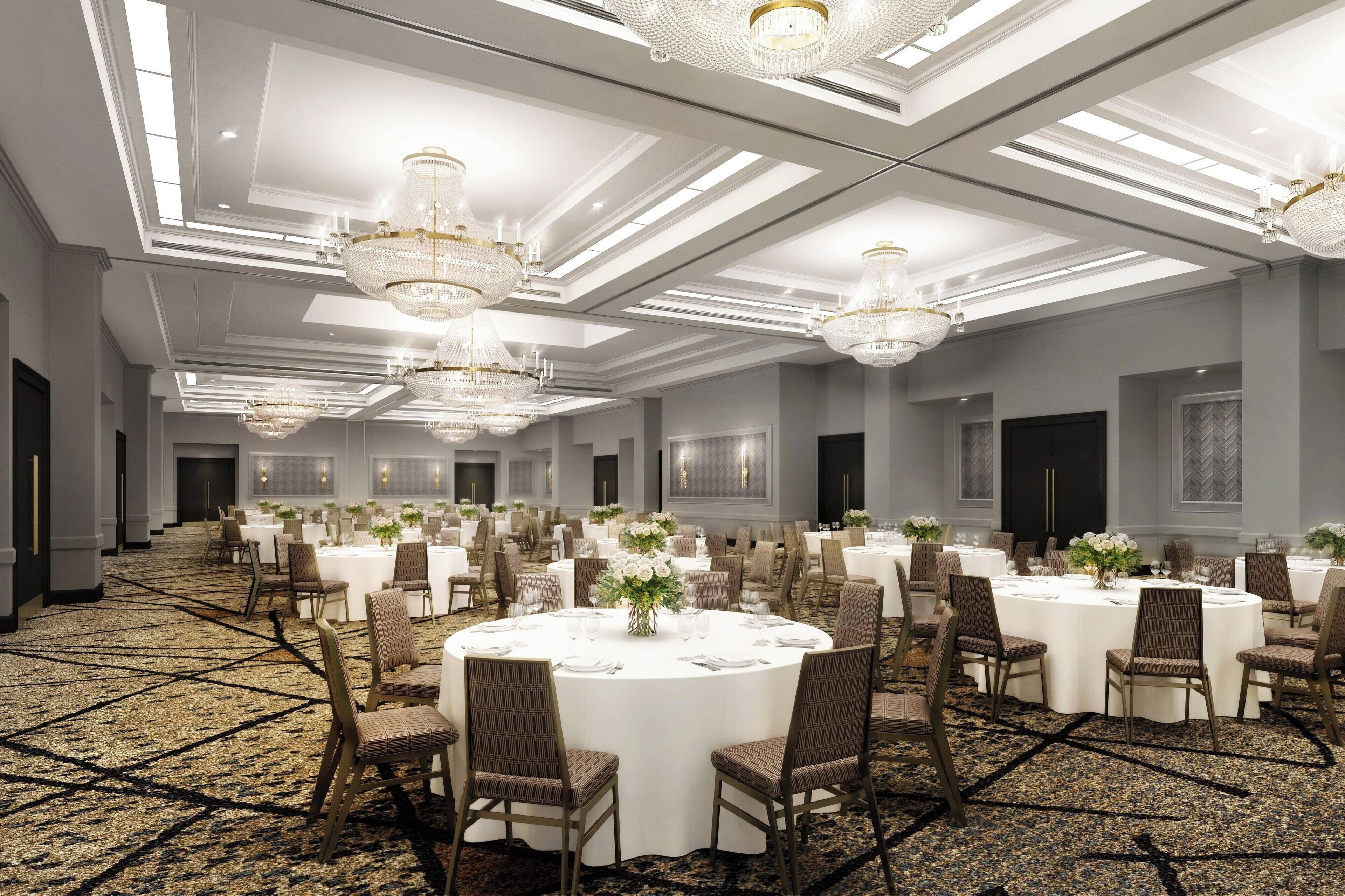 A large, well-lit banquet hall with round tables, elegant chandeliers, and a carpeted floor.