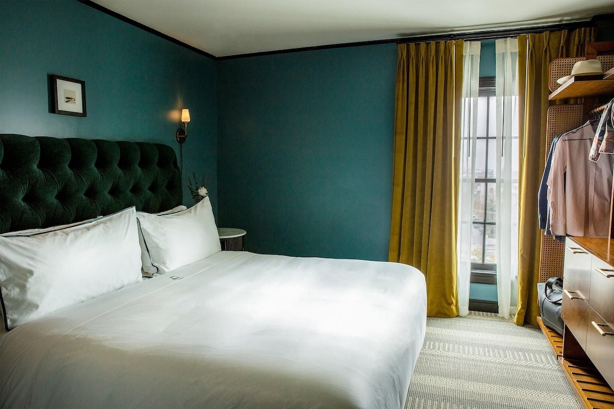 Habitación de hotel con una cama grande, una mesita de noche, una lámpara, un armario y una ventana con cortinas.