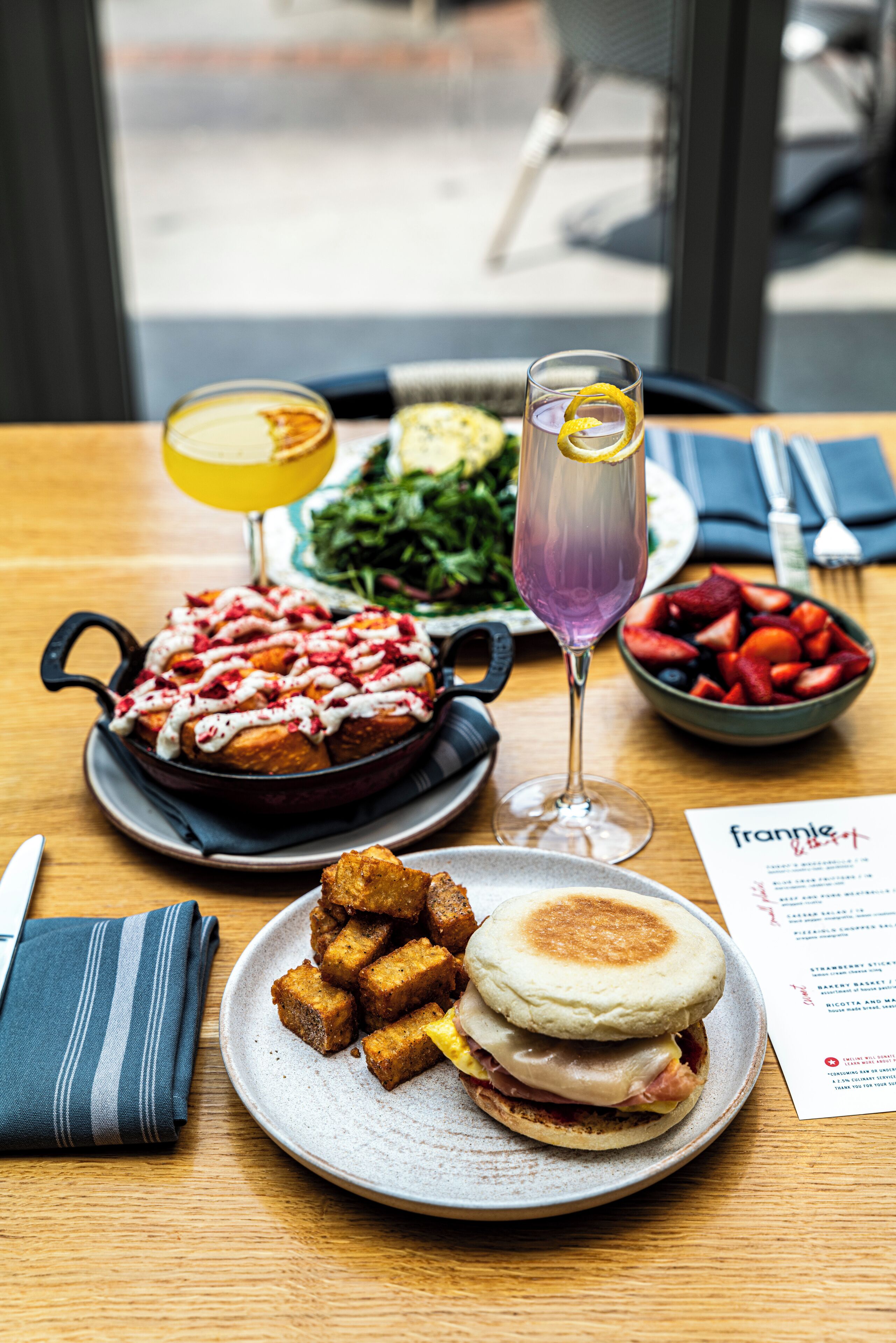 A dining table with a meal consisting of a breakfast sandwich, a skillet with pancakes, a bowl of fruit, and two drinks.