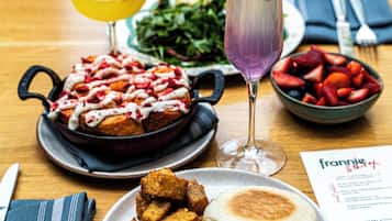 A dining table with a meal consisting of a breakfast sandwich, a skillet with pancakes, a bowl of fruit, and two drinks.