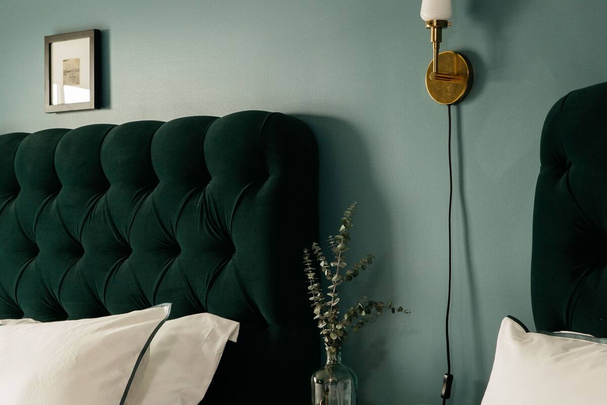 A bedroom with a tufted headboard, a bedside table with a vase and books, and a wall-mounted lamp.