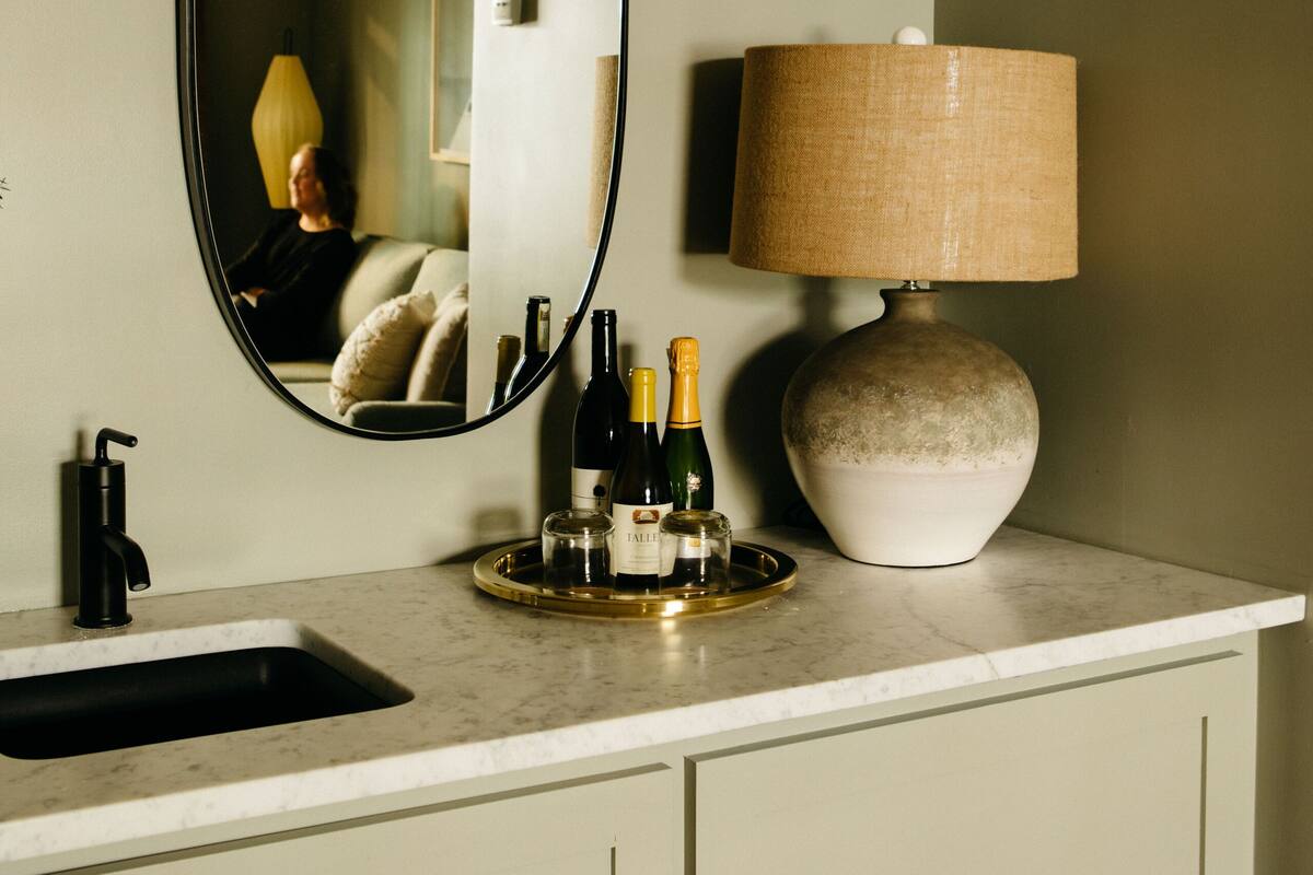 A bathroom vanity with a mirror, a sink, a bottle of champagne, and a lamp.