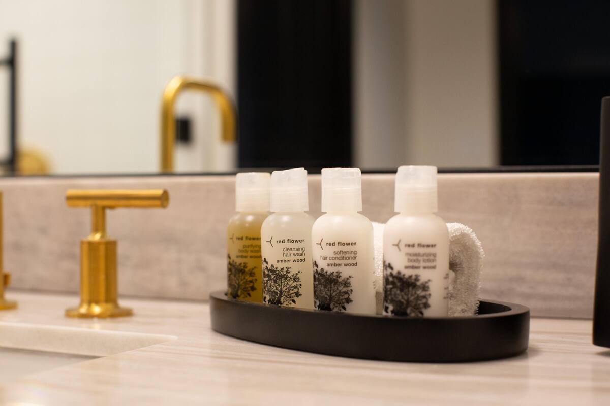 A bathroom with a black tray holding four bottles of red flower body lotion and a gold faucet.
