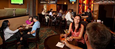 A bar area with multiple seating arrangements, a television mounted on the wall, and a bar stocked with various bottles.