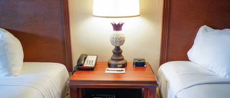 A hotel room with two beds, a bedside table with a lamp, a telephone, and a digital clock.