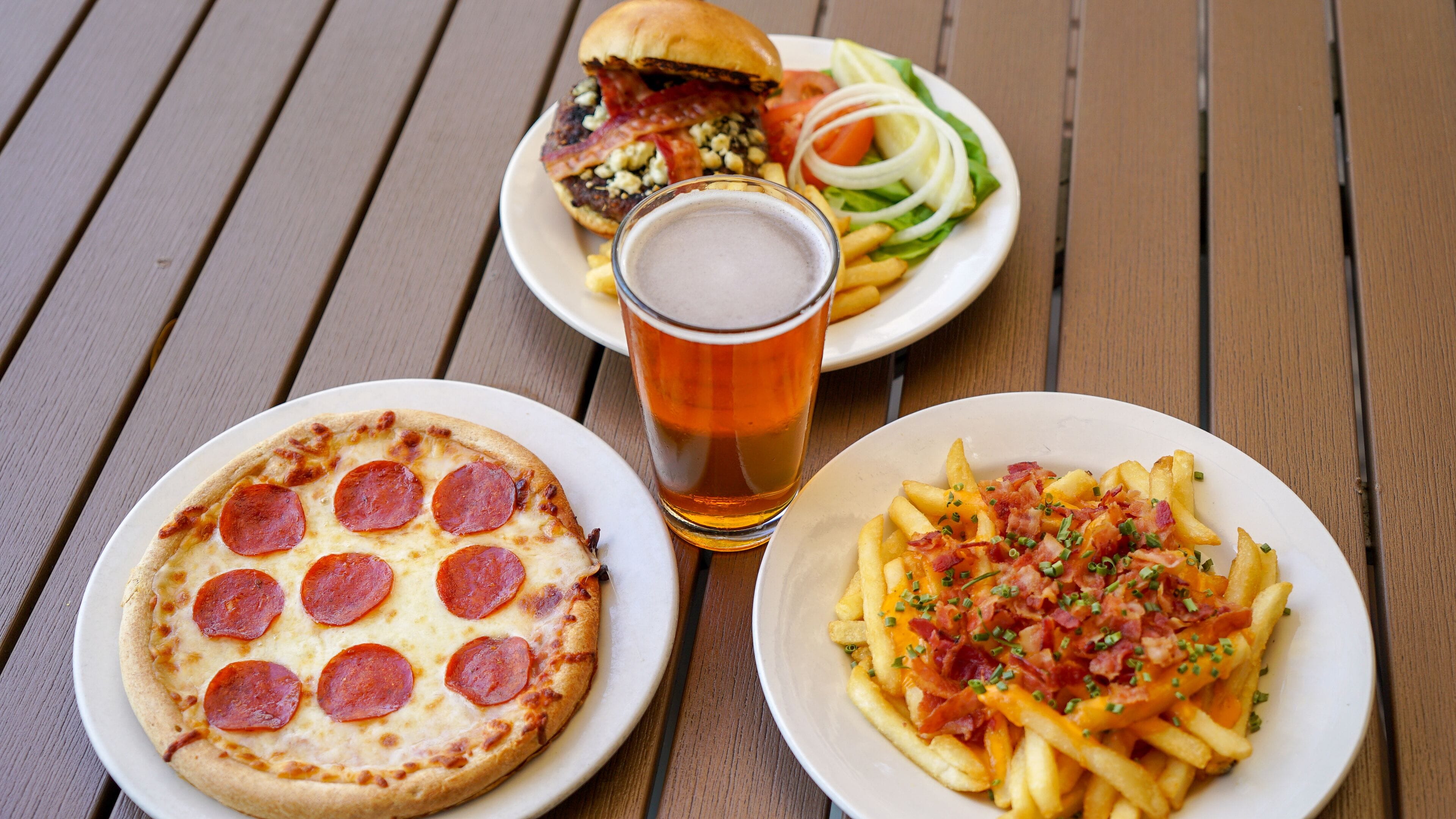 A table with three plates of food: a pepperoni pizza, a burger with bacon, and a plate of fries topped with bacon and cheese.