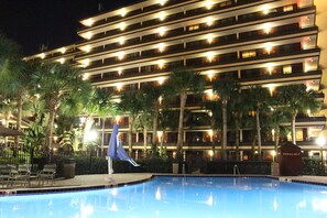 A hotel with a swimming pool, palm trees, and a well-lit exterior.