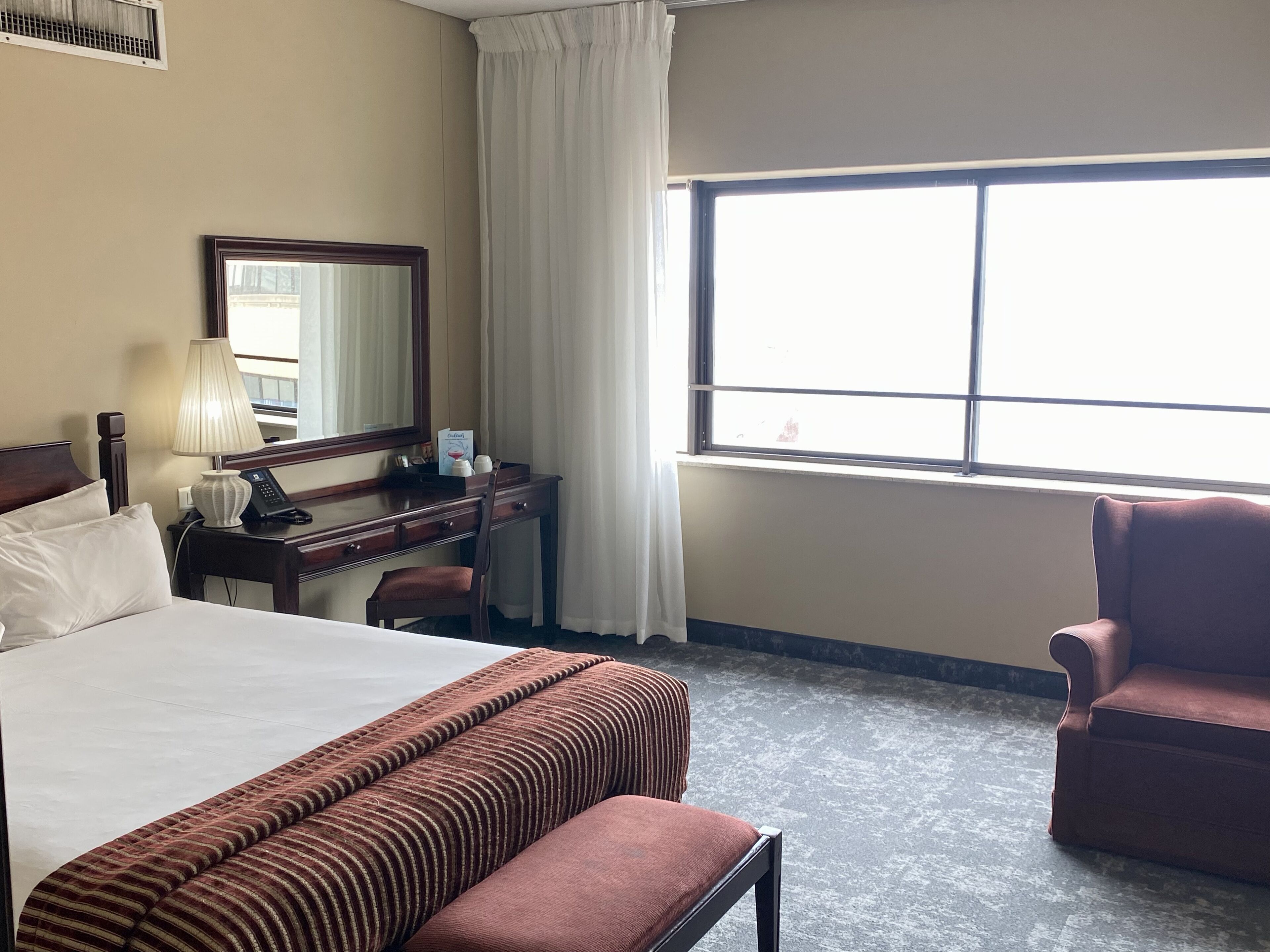 A hotel room with a bed, a desk with a lamp, a chair, and a large window.