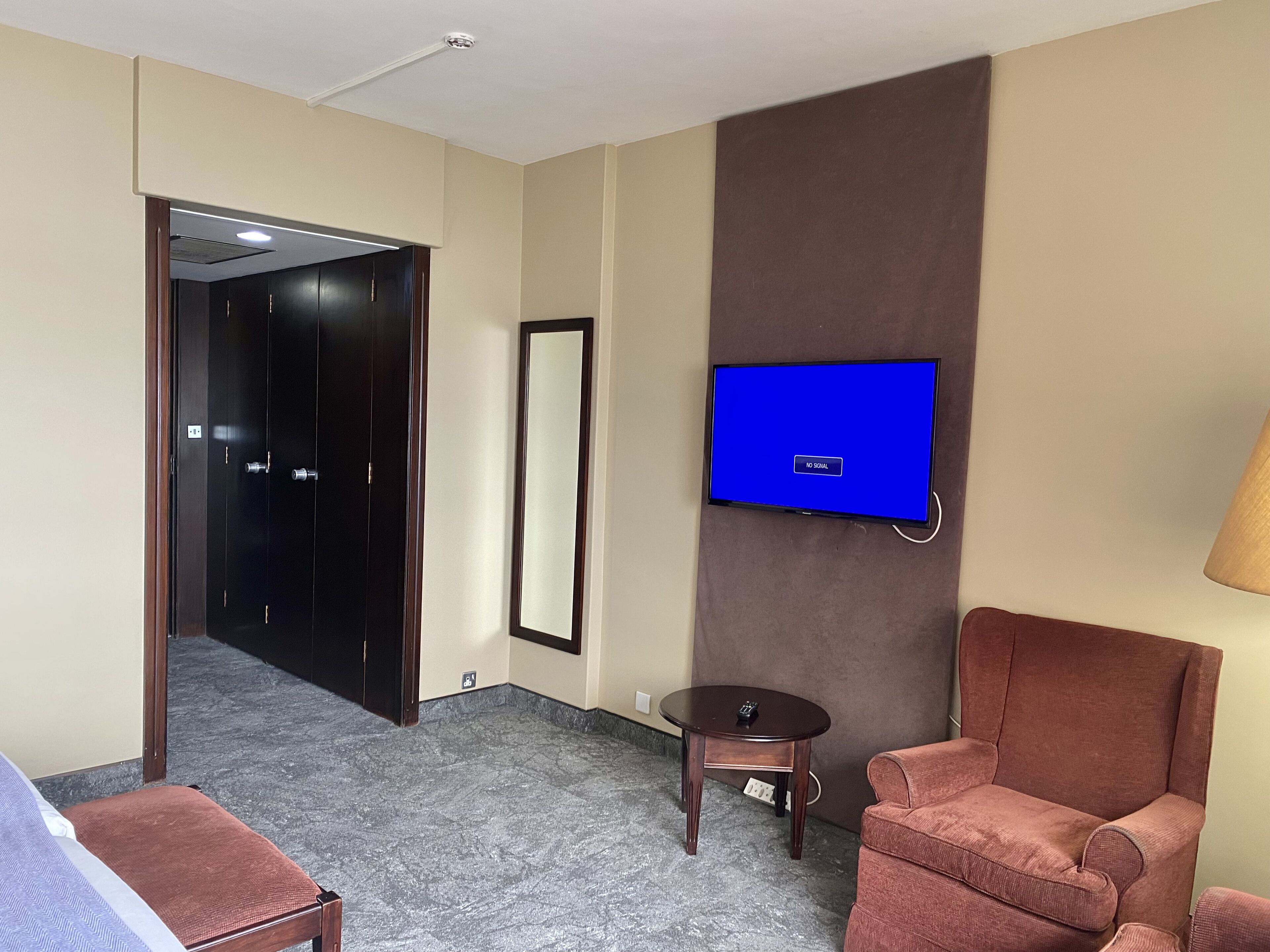 A hotel room with a flat-screen TV mounted on the wall, a brown armchair, a small round table, and a bed with a blue and white striped bedspread.