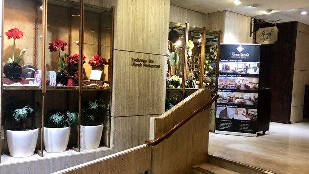 Zona de recepción de un hotel con una escalera de madera, plantas en macetas y un cartel que anuncia el bar Exchange y el restaurante Ulundi.