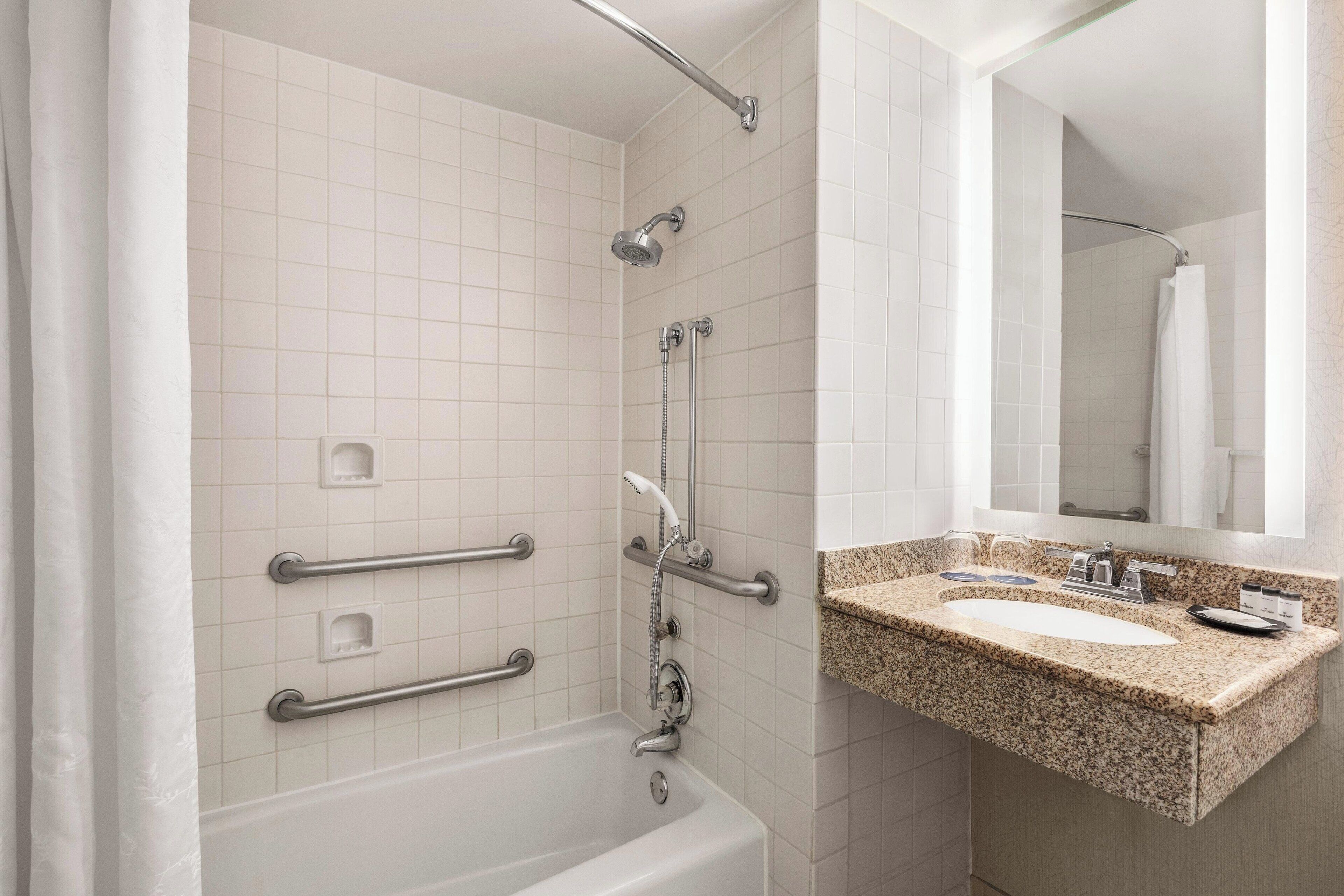 Un baño con una bañera blanca, barras de apoyo, cabezal de ducha, un lavamanos con encimera de granito y un espejo.