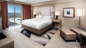A modern hotel room with a large bed, a desk, a chair, and a view of the city.