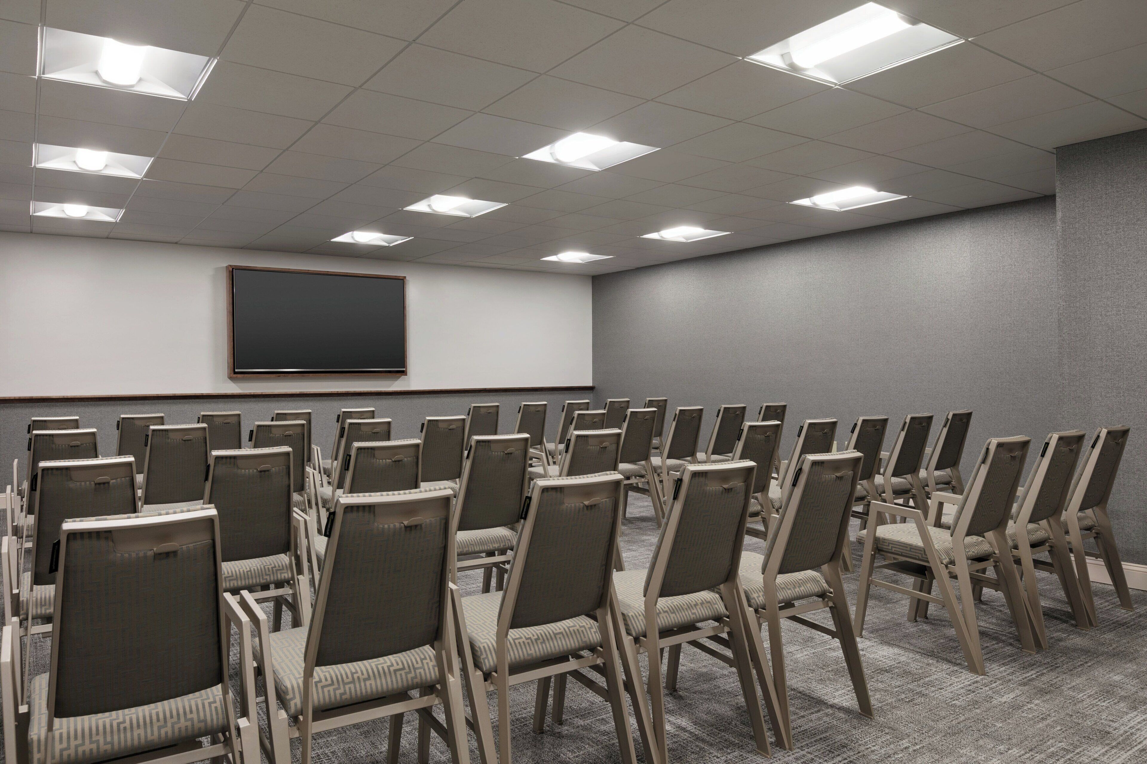 Une salle de conférence avec des rangées de chaises, un grand téléviseur à écran plat fixé au mur et des luminaires au plafond.
