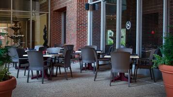 An outdoor seating area with brick walls and large windows, featuring wicker chairs and tables.