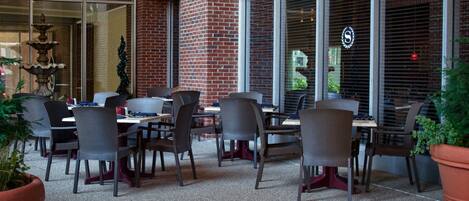 An outdoor seating area with brick walls and large windows, featuring wicker chairs and tables.