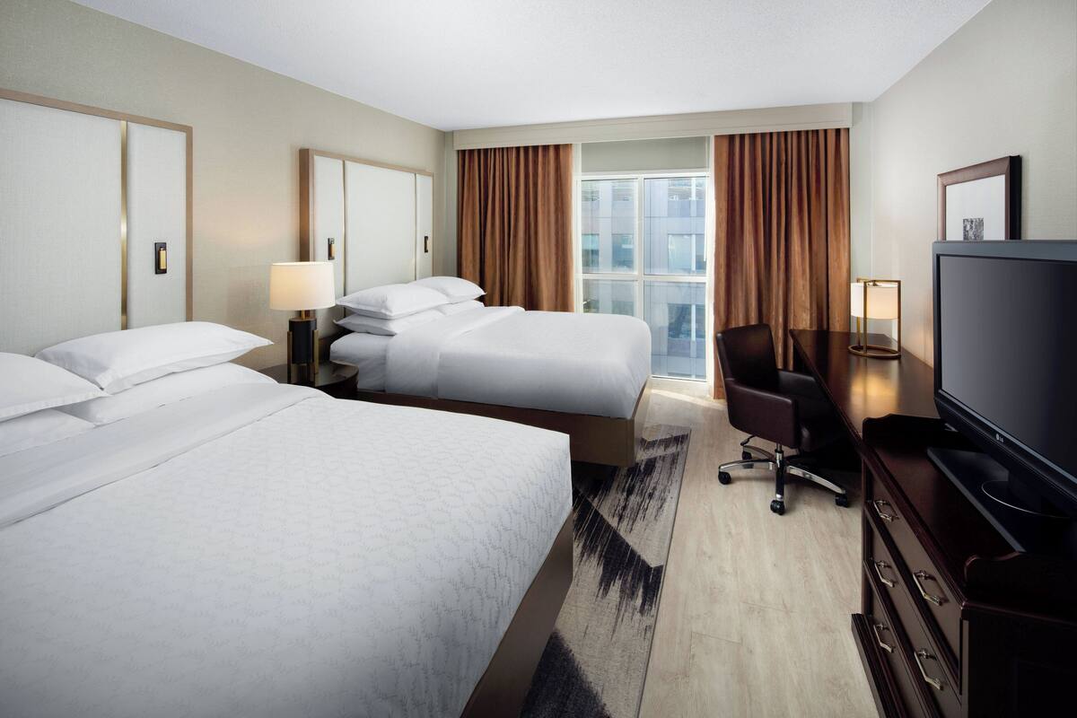 Quarto de hotel com duas camas, uma escrivaninha, uma cadeira, televisão e uma janela grande com cortinas.
