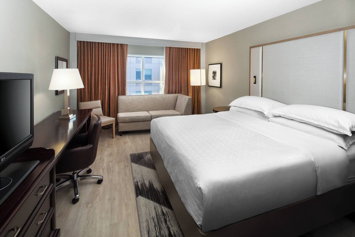 Quarto de hotel com cama, escrivaninha com televisão, sofá e janela com cortinas.