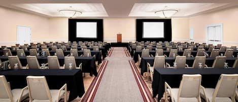 A conference room with rows of tables and chairs, a carpeted aisle, and a projector screen at the front.