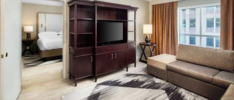 A hotel room with a large flat-screen TV, a sofa, a wooden entertainment unit, a bed with white linens, and a view of the city through a large window.