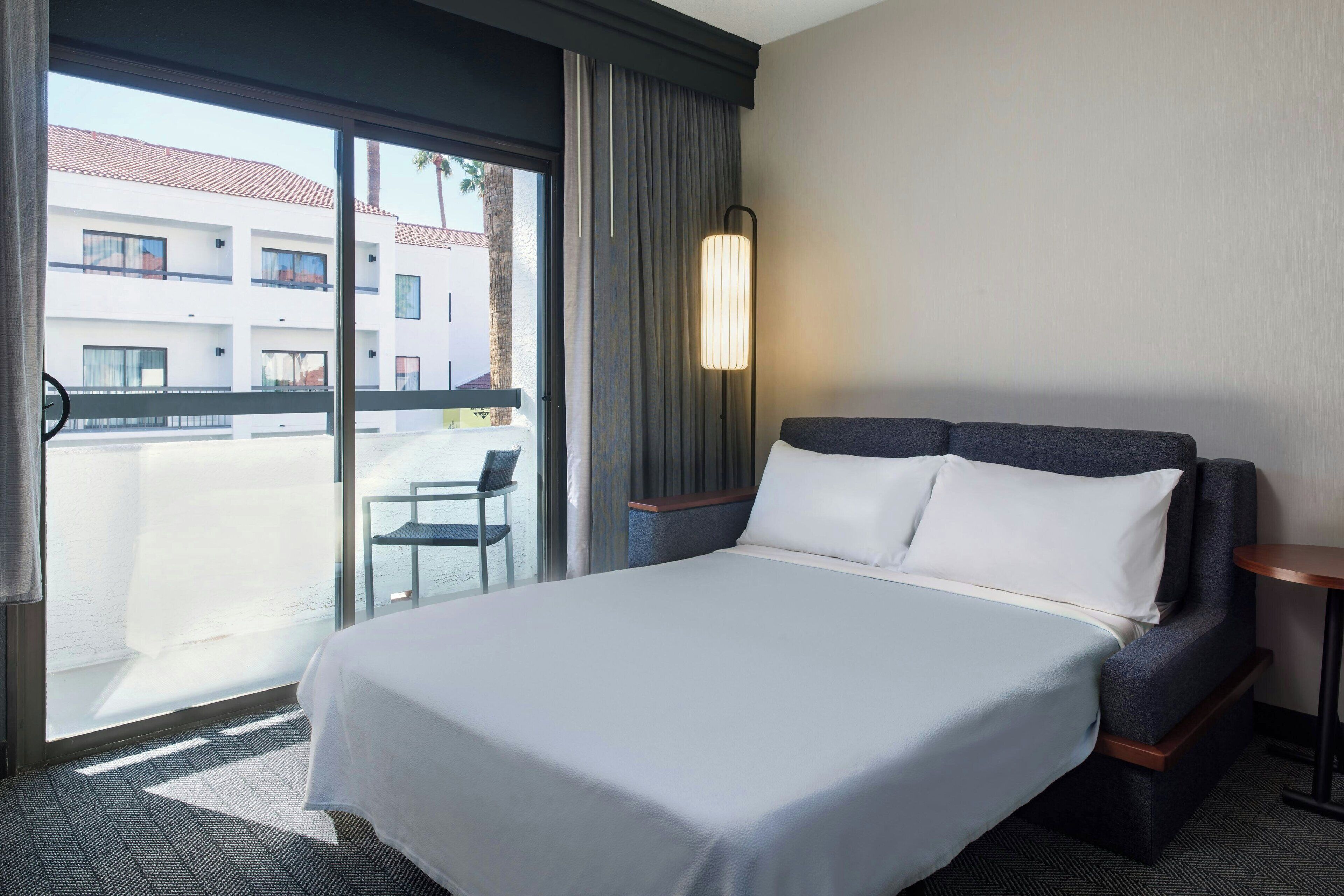 Een hotelkamer met een groot bed, een kleine tafel, een stoel en uitzicht op een gebouw door een glazen deur.