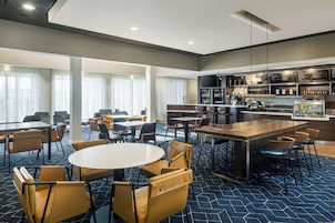 A hotel lobby with round tables and chairs, a bar area, and a seating area with large windows.