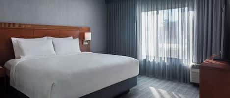 Une chambre d’hôtel avec un grand lit, une tête de lit en bois, une table de chevet, un bureau et une fenêtre avec des rideaux.