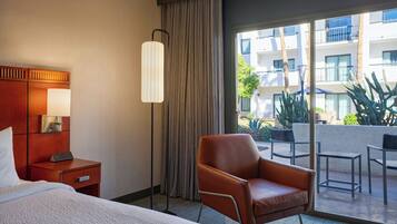A hotel room with a bed, a nightstand, a lamp, a chair, and a view of the outdoor balcony with plants and a building in the background.