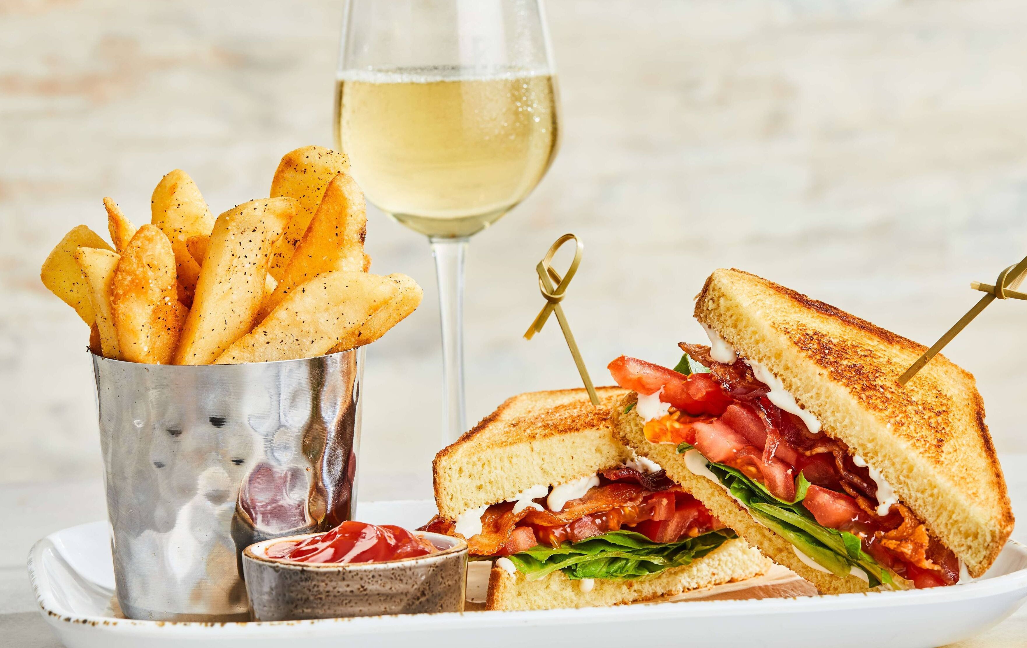 A sandwich with bacon, tomato, and lettuce, a glass of white wine, and a container of seasoned fries.