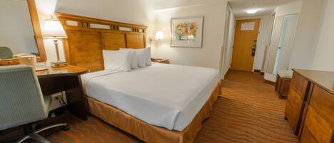 A hotel room with a large bed, a desk with a chair, a desk lamp, a mirror, and a painting on the wall.
