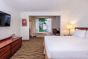 Habitación de hotel con cama, escritorio, silla, televisor, lámpara y ventana con cortinas.