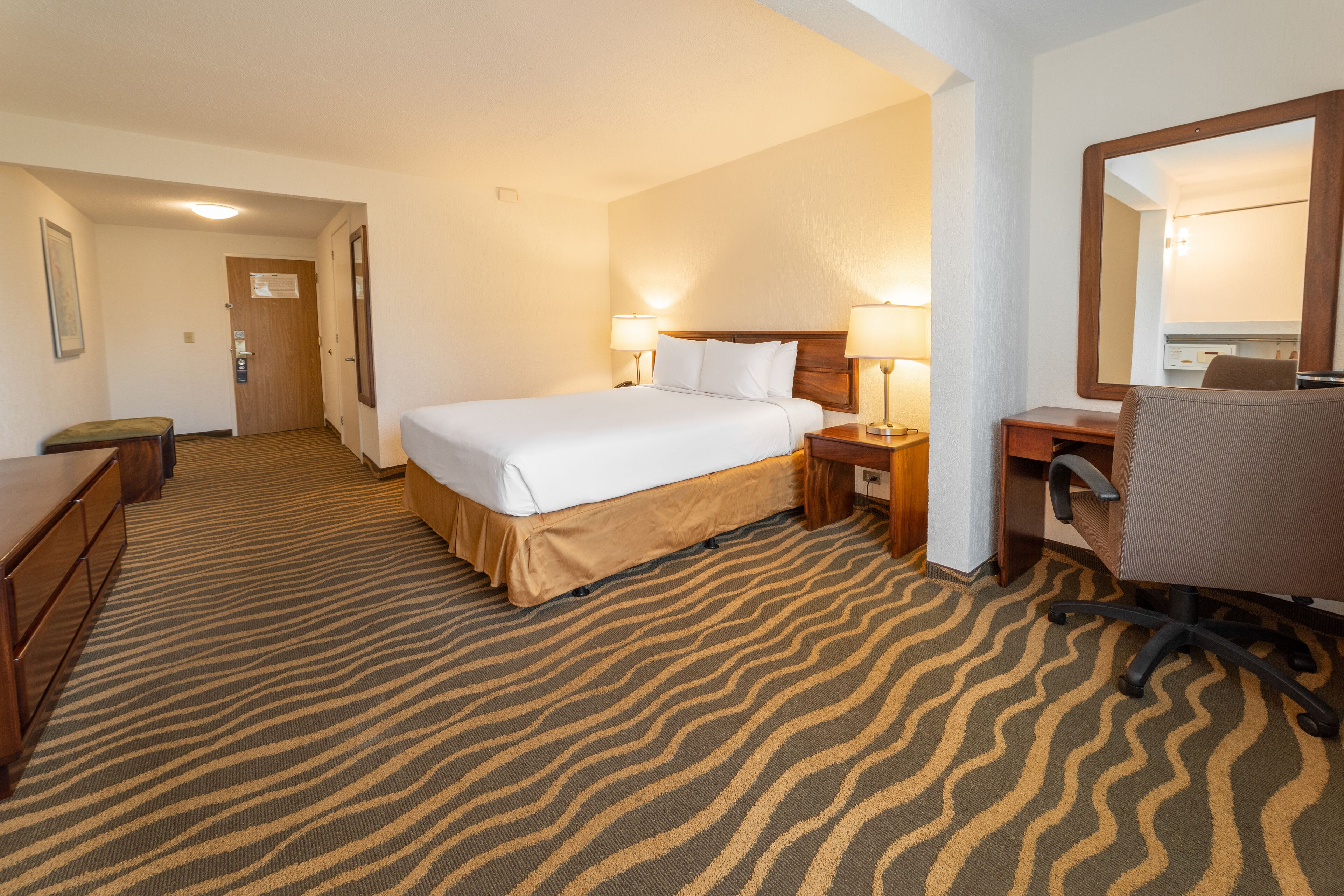 Ein Hotelzimmer mit einem Bett, Nachttischen, einem Schreibtisch, einem Sessel und einem Spiegel.
