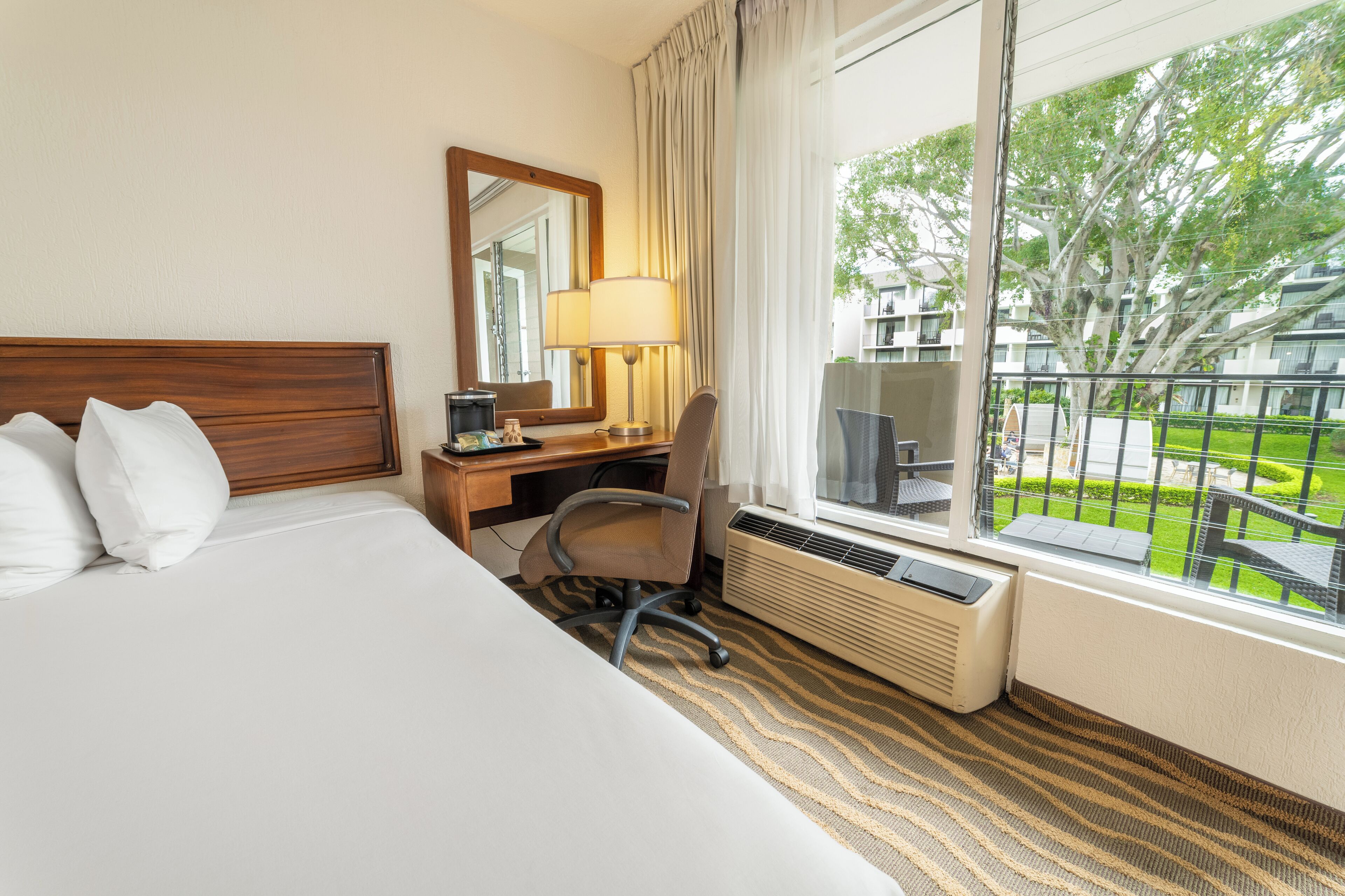 A hotel room with a bed, desk, chair, and a large window with a view of the outdoors.