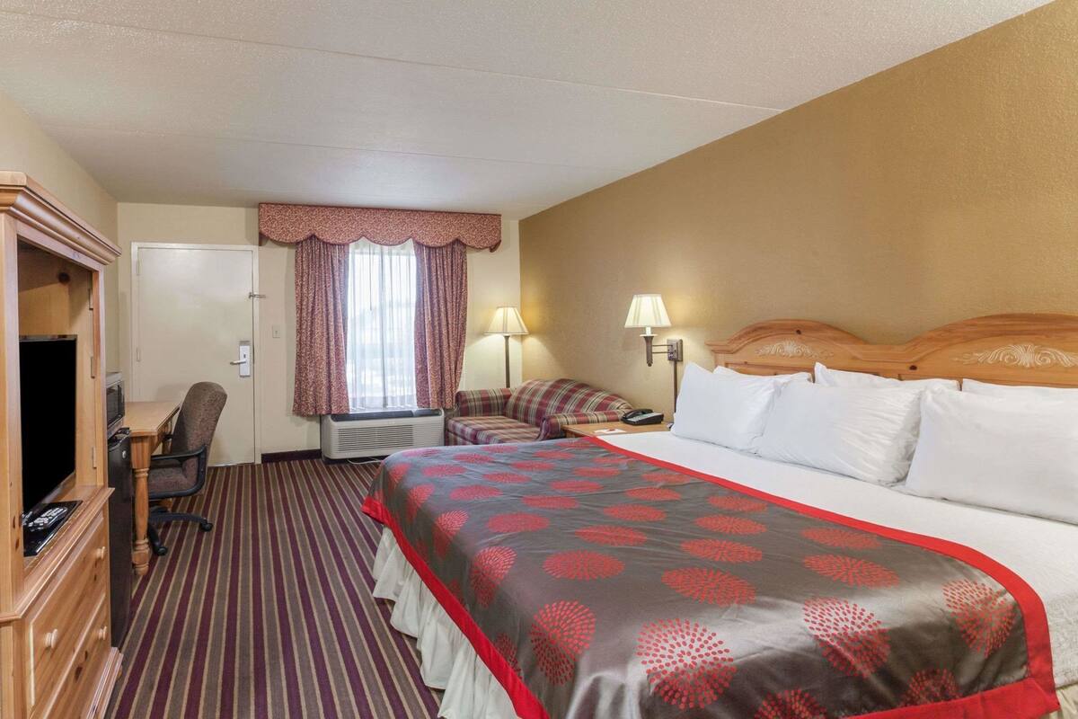 A hotel room with a bed, a desk with a chair, a television, a window with curtains, and a lamp.