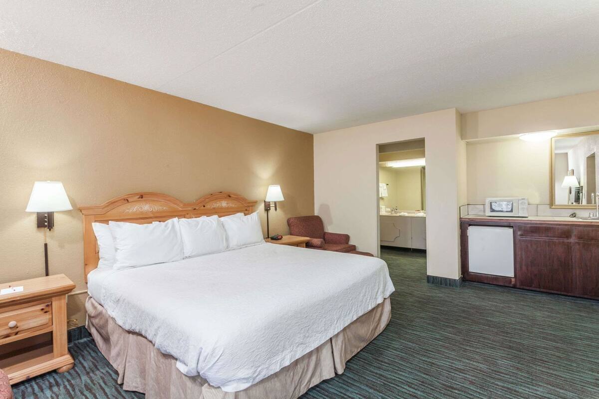 A hotel room with a large bed, bedside lamps, a chair, a desk, and a small kitchenette.