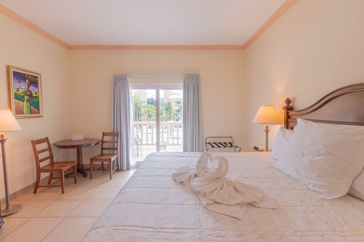 Quarto de hotel com cama, escrivaninha, duas cadeiras, luminária e um quadro na parede.