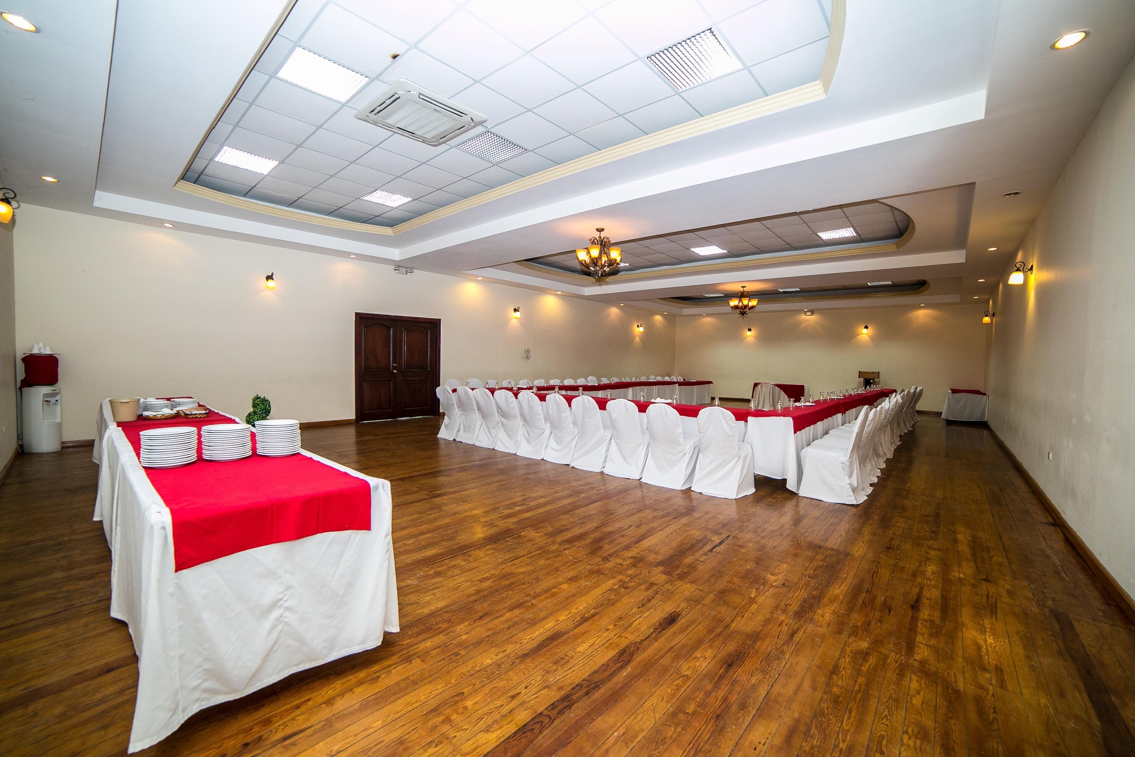 Une grande salle de conférence avec une longue table recouverte d’une nappe rouge, des chaises blanches et un plancher en bois.