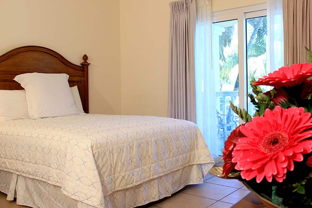 Uma cama com cabeceira de madeira, edredom branco e uma grande flor vermelha em um vaso.