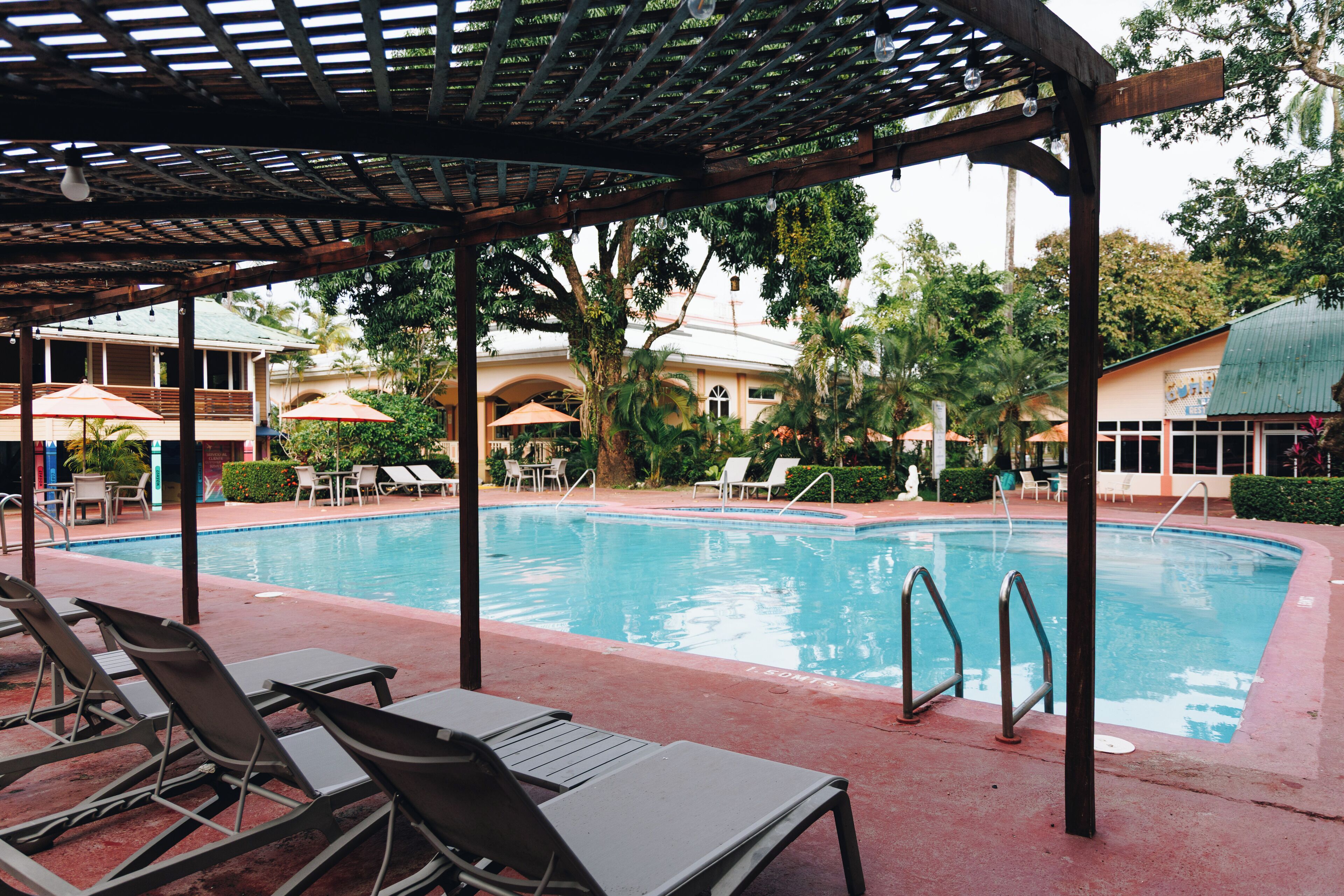 A pool area with lounge chairs and umbrellas, surrounded by greenery and buildings.