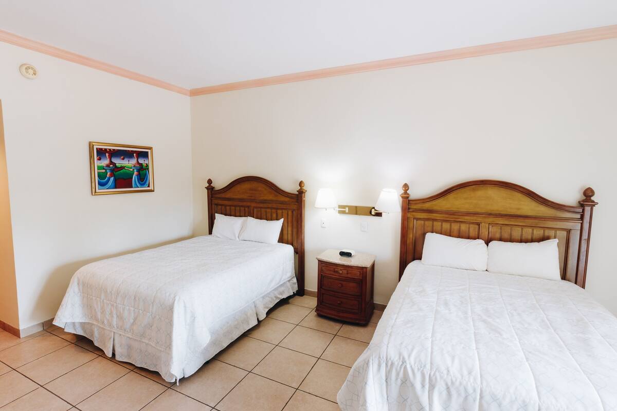 Quarto de hotel com duas camas de solteiro, uma mesa de cabeceira com abajur e um quadro na parede.